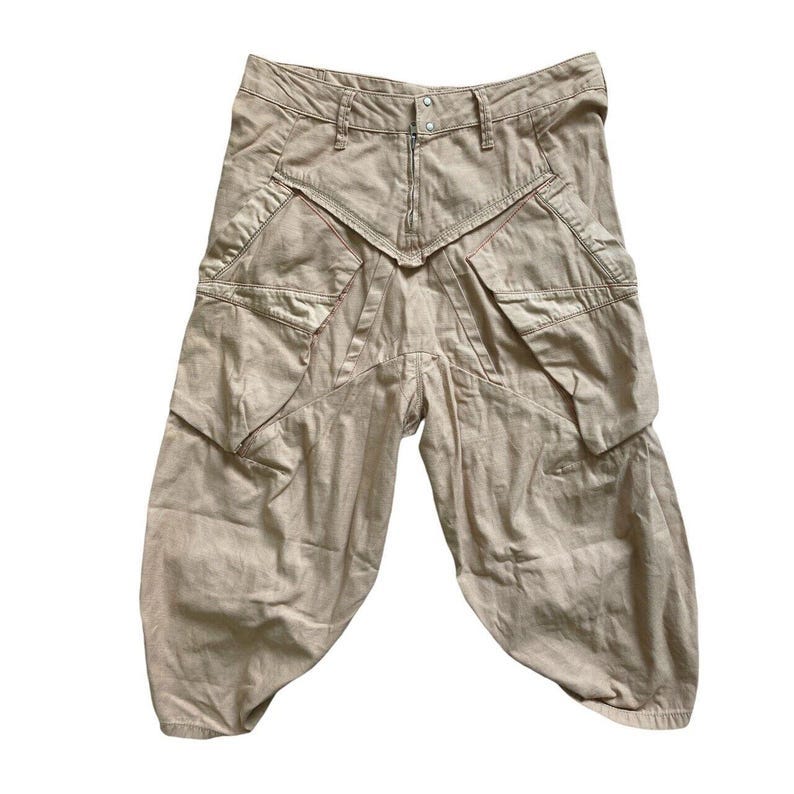 May include: Beige cargo pants with multiple pockets and a unique design. The pants have a wide leg and a relaxed fit.