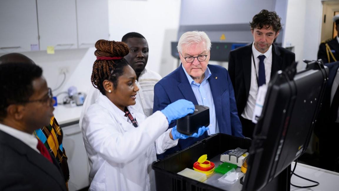 Eine Gruppe von Personen, darunter ein prominenter Gast, beobachtet eine Wissenschaftlerin, die ein wissenschaftliches Gerät im Labor vorführt.