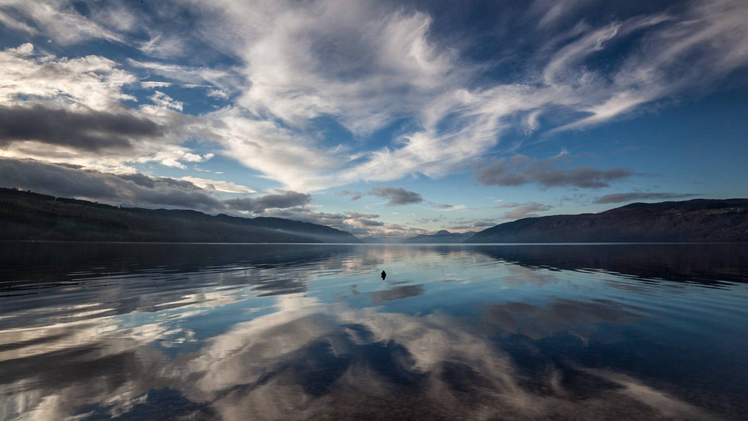 Wide view of calm Loch Ness with cloud reflections; low hills on both sides, small dark buoy/stone near centre.