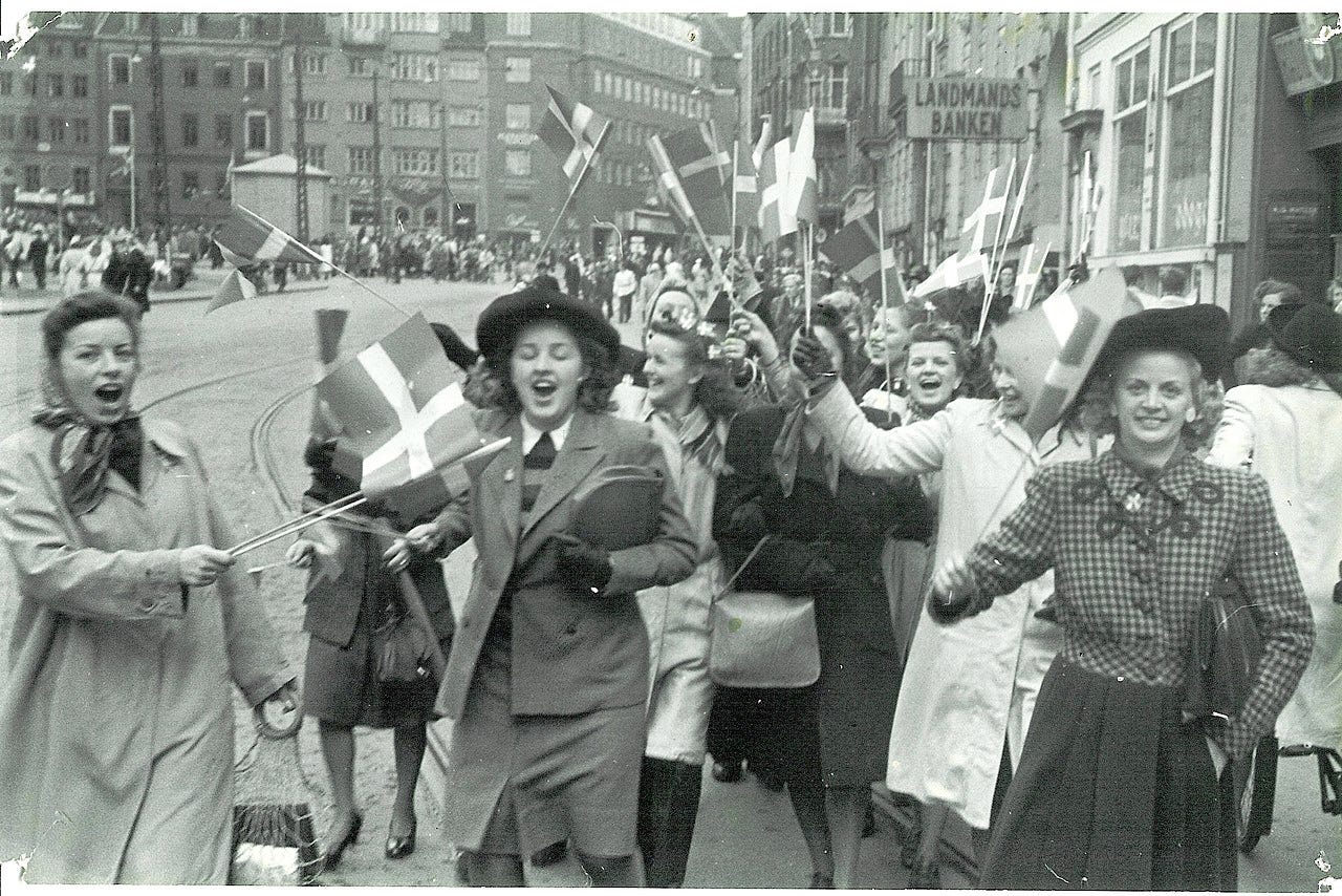 Young women cheering and smiling waving Danish flags as they walk down a broad city street