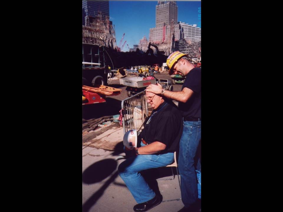 Chiropractor in a yellow hard hat adjusts a seated man's neck with cranes and rubble at Ground Zero behind them