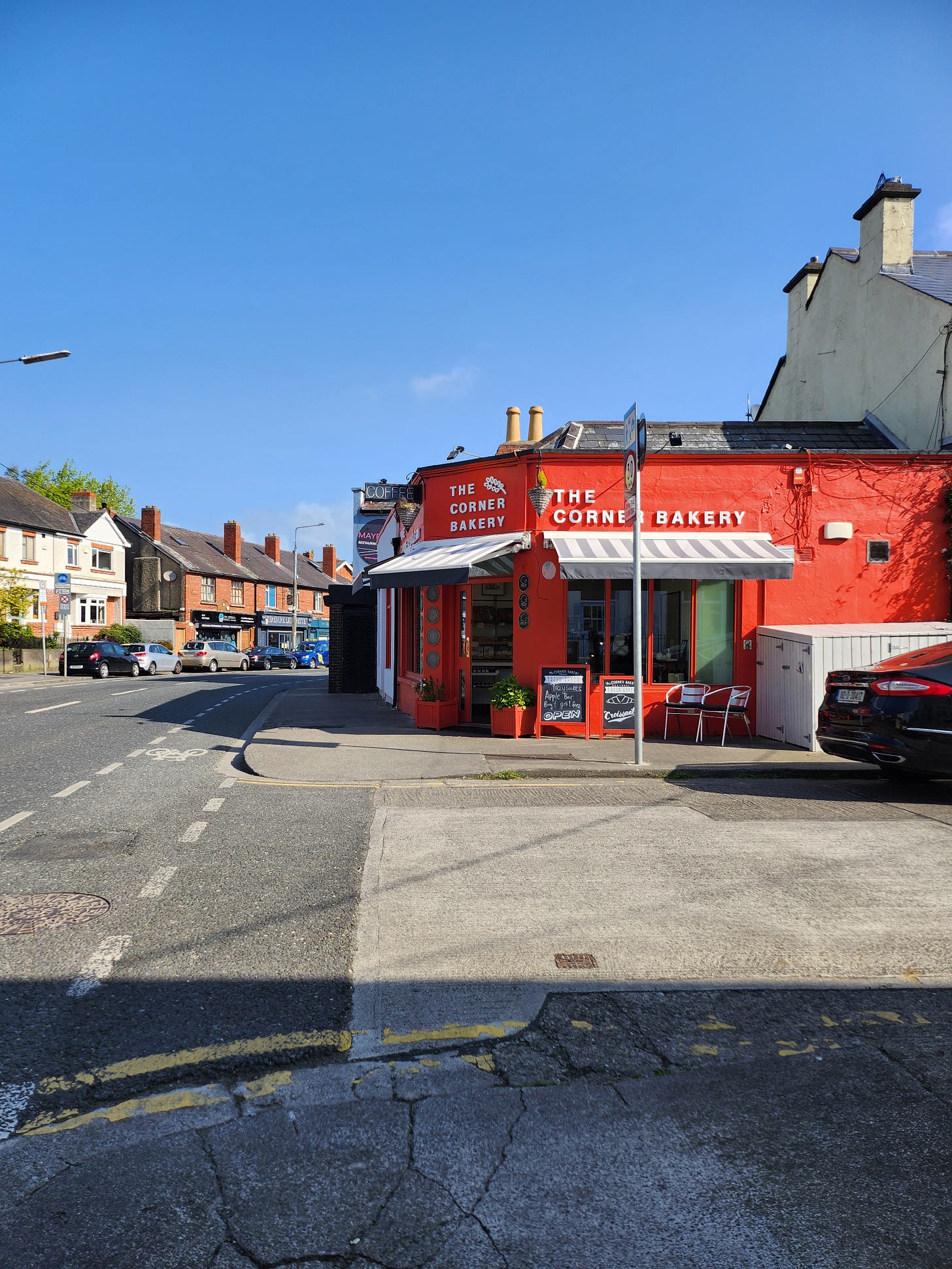 The Corner Bakery, a bright red bakery sits on the corner of a main road and a housing cul de sac. Bright blue skies fall behind. 