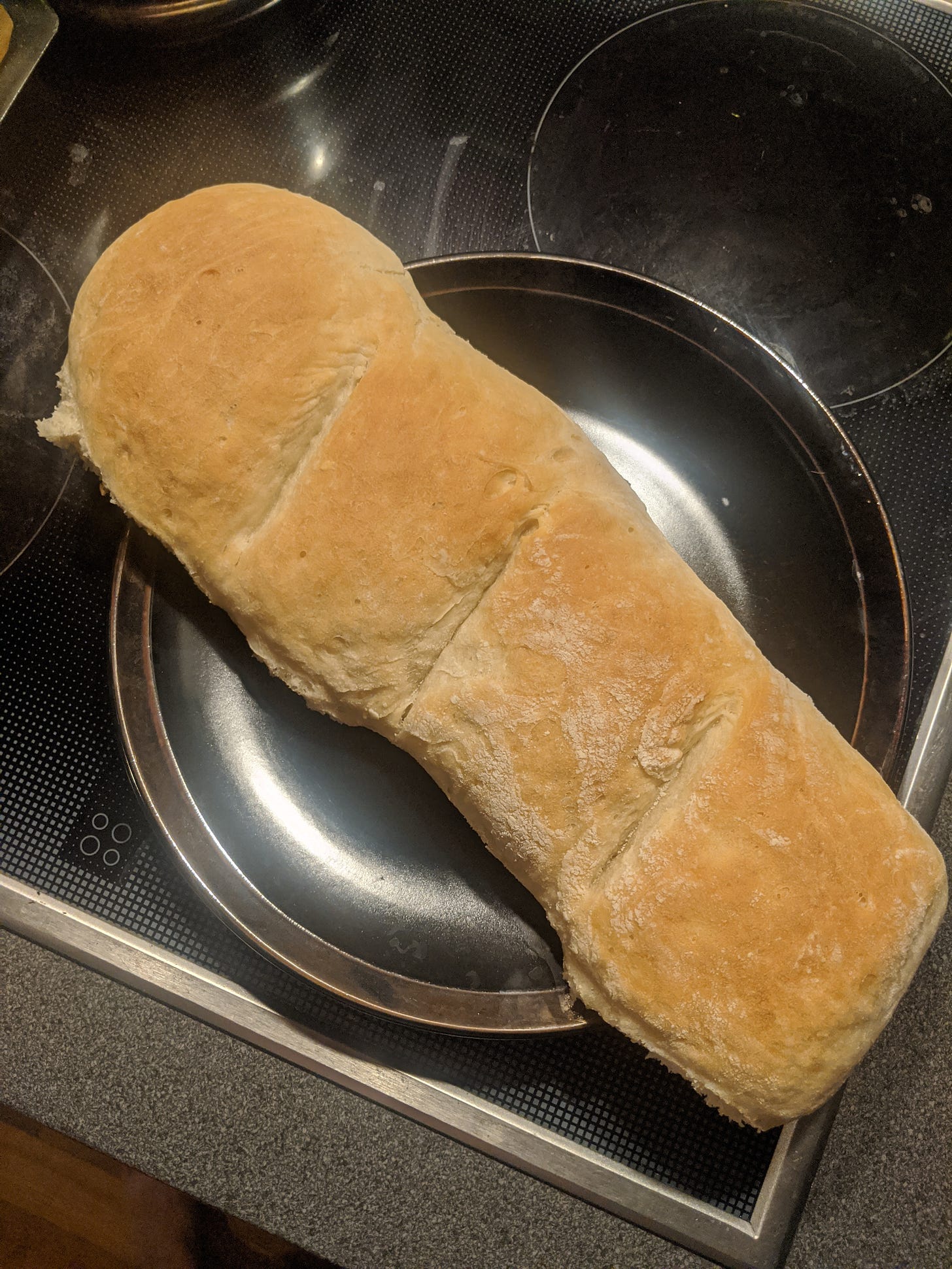 A loaf of French bread, in the US-American definition, with a somewhat soft, golden exterior. About 1.5 feet or a half-meter long, with 3 light scores on the top, set atop a black plate.