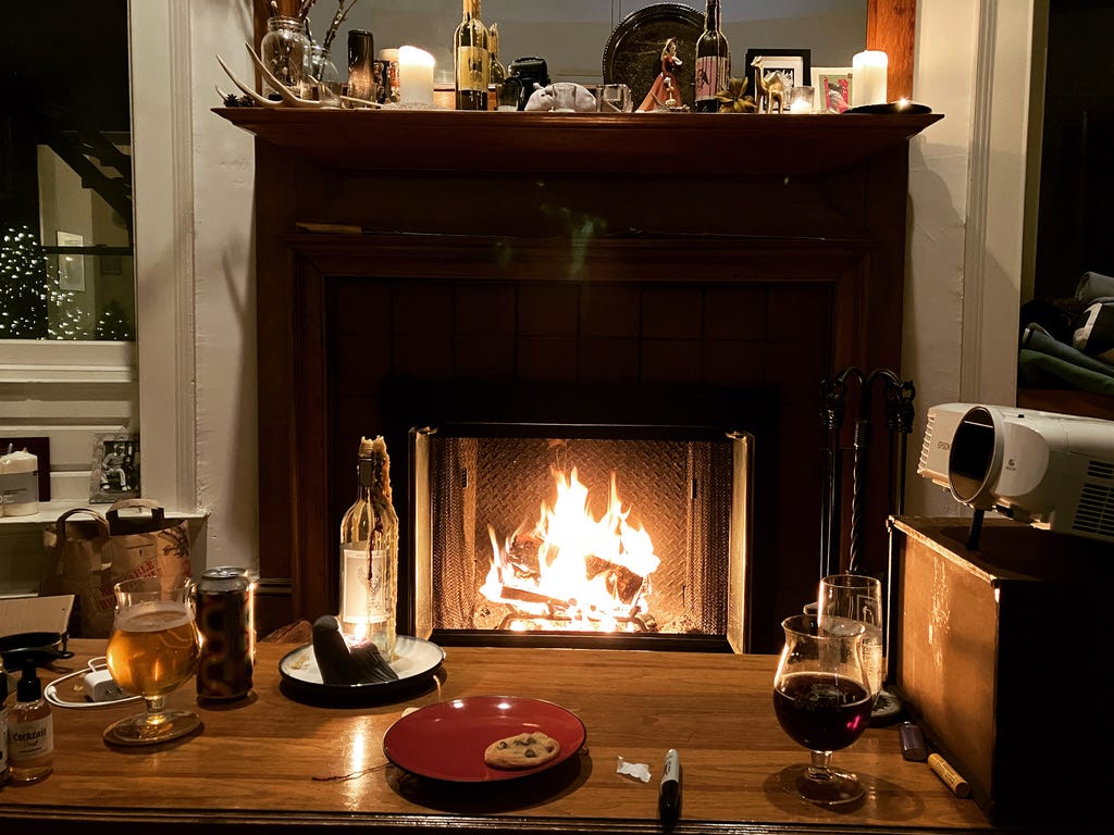 a still life of a roaring fire place, lit candles, two glasses of beer, a single cookie remaining on a plate, and other knic knacks decorating the mantle place and coffee table.