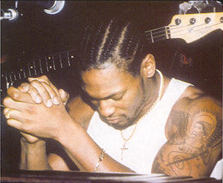 D’Angelo, with his hair in neat braids and wearing a white tank top, is seated in front of a bass guitar; his arms are folded and resting on a table, showing his prominent tattoo, as his head is bowed and his face is partially hidden. D’Angelo, with his hair in neat braids and wearing a white tank top, is seated in front of a bass guitar; his arms are folded and resting on a table, showing his prominent tattoo, as his head is bowed and his face is partially hidden.