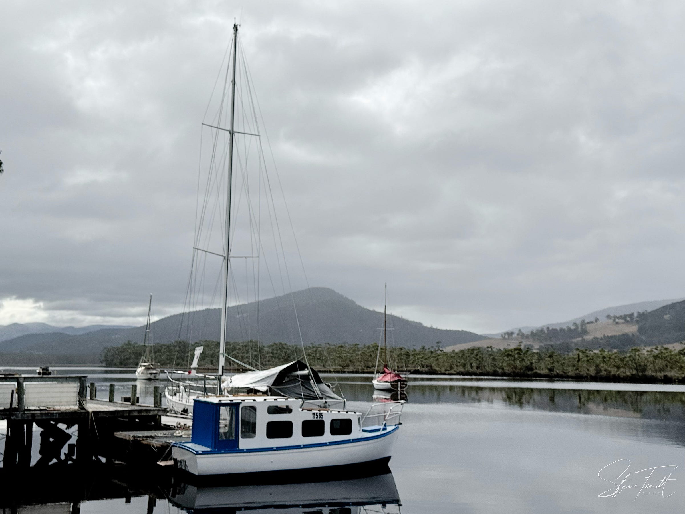 Photograph: a wooden jetty on a wide river with a triangular wooded peak in the background. Traditional yachts and a small cabin cruiser alongside the jetty.