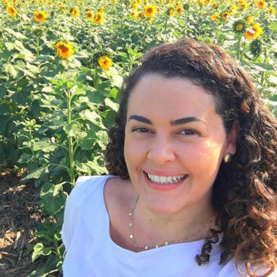 Imagem de uma mulher branca de cabelos pretos e longos. Sorri e veste uma blusa branca. Fundo, campo de flores.