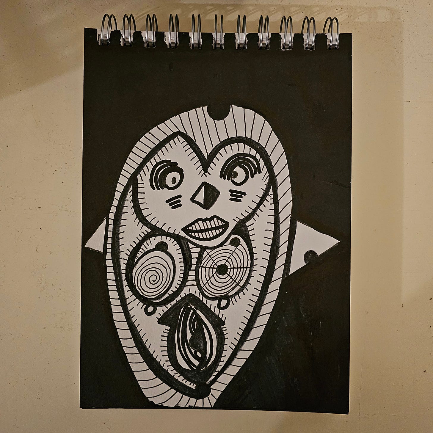  * A high-contrast black and white ink drawing of a stylized creature. The figure has a heart-shaped face, large hypnotic eyes (one spiral, one concentric), and patterns resembling a penguin or owl. The background is filled with solid black ink.