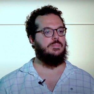 A imagem contém um homem com cabelo e barba escura, ele usa óculos