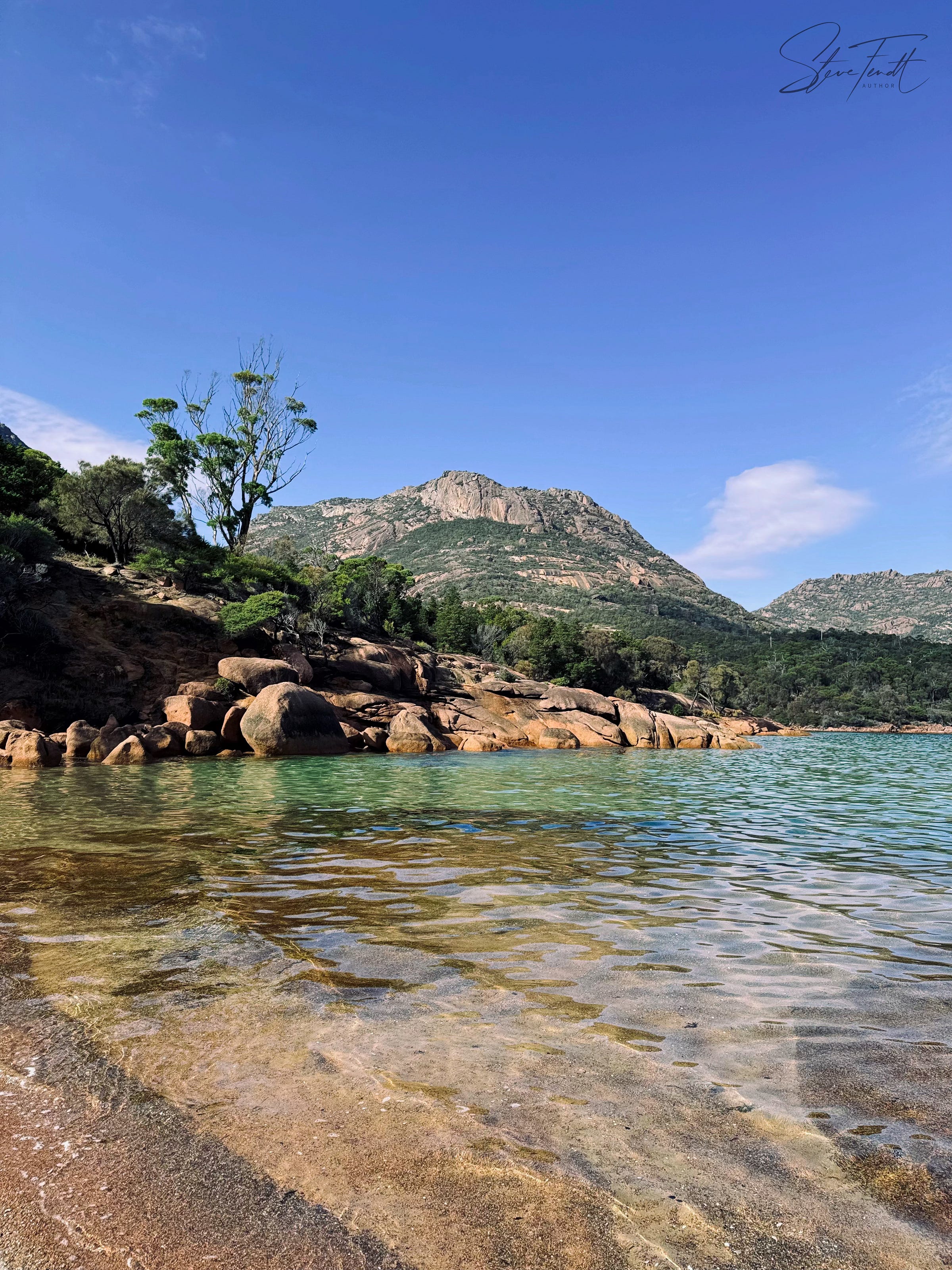 Beachscape: a small beach between rounded, reddish rocks, dark green bushes, in the distance a rugged peak, under blue skies. The water is crystal clear and shallow.