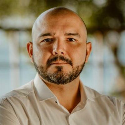 Homem calvo e de barba, usando camisa branca, tendo ao fundo uma janela e uma árvore Homem calvo e de barba, usando camisa branca, tendo ao fundo uma janela e uma árvore
