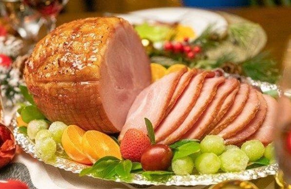 A glazed ham partially sliced on a decorative silver platter, garnished with fresh fruits and greenery.