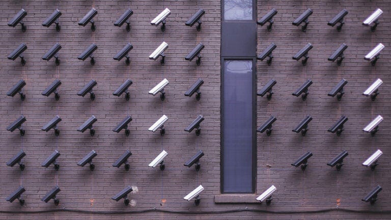 Rows of security cameras on a brick wall beside a tall, narrow window.