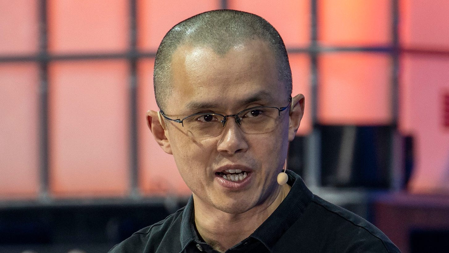 Changpeng Zhao looking toward and the left of the camera, speaking to an audience it a small microphone extending from his ear.