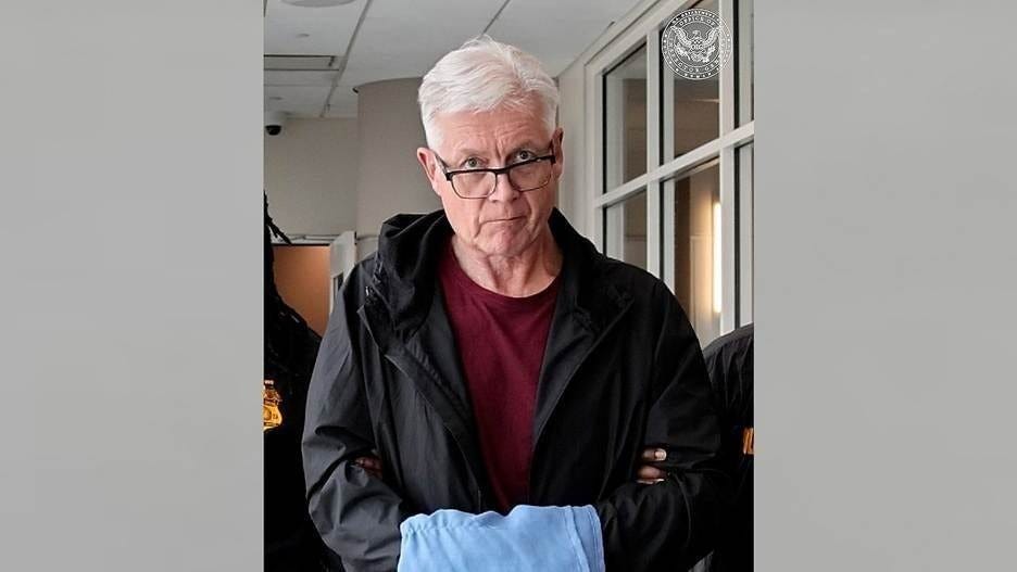 Older man with white hair and glasses, in a dark jacket over a maroon shirt, escorted by authorities in a federal office. 