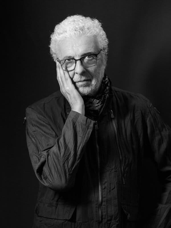 A black-and-white portrait of André Heller, in glasses, with his right hand resting on his cheek.