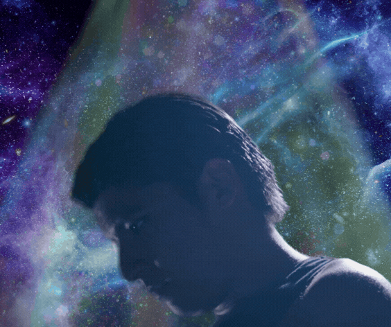 In front of a blueish-purple background of nebulas and stars, a man slowly lifts his head in profile. He is in the center of a subtle spotlight with falling transparent orbs and star-like shapes.