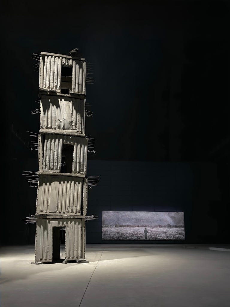 Installation view of Anselm Kiefer’s The Seven Heavenly Palaces, showing a tall, precarious tower in a dark exhibition space. The work emphasizes scale, weight, verticality and architectural instability.