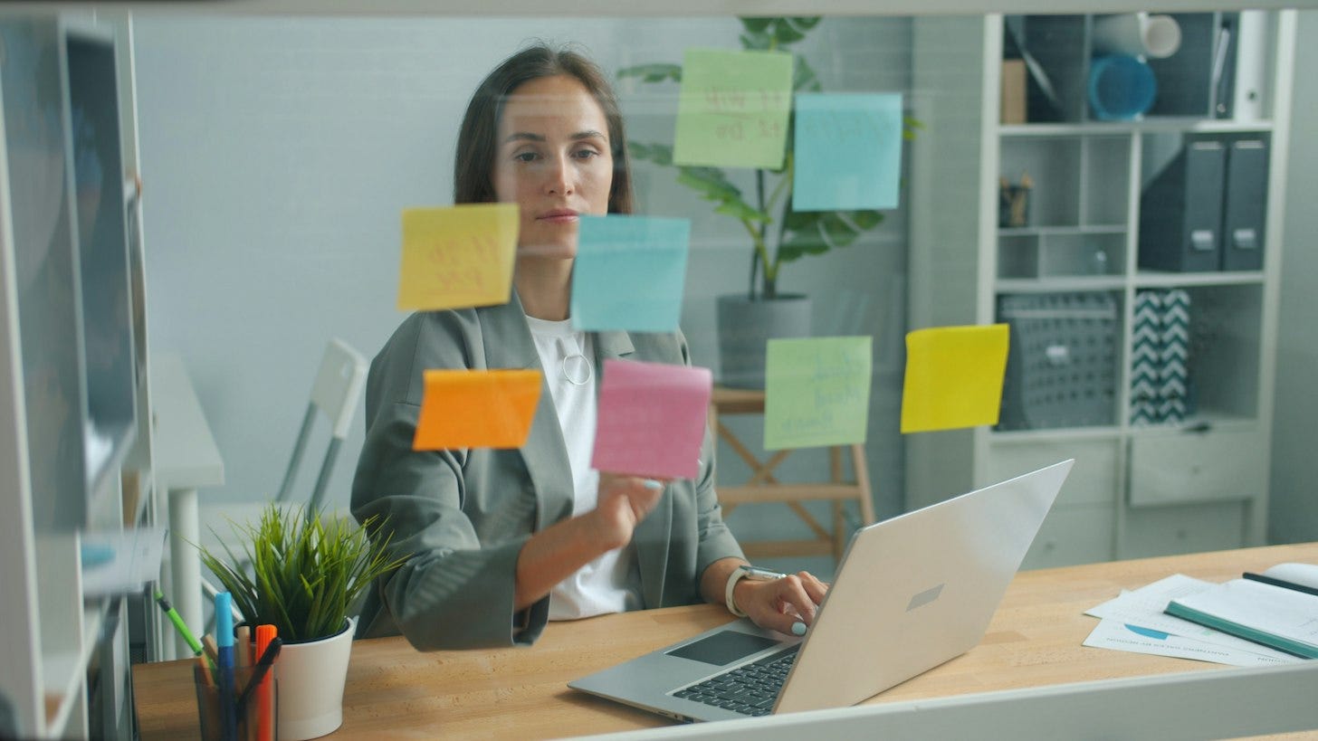 A woman at a desk with an open laptop sticks colorful notes onto a glass board in front of her. A woman at a desk with an open laptop sticks colorful notes onto a glass board in front of her.