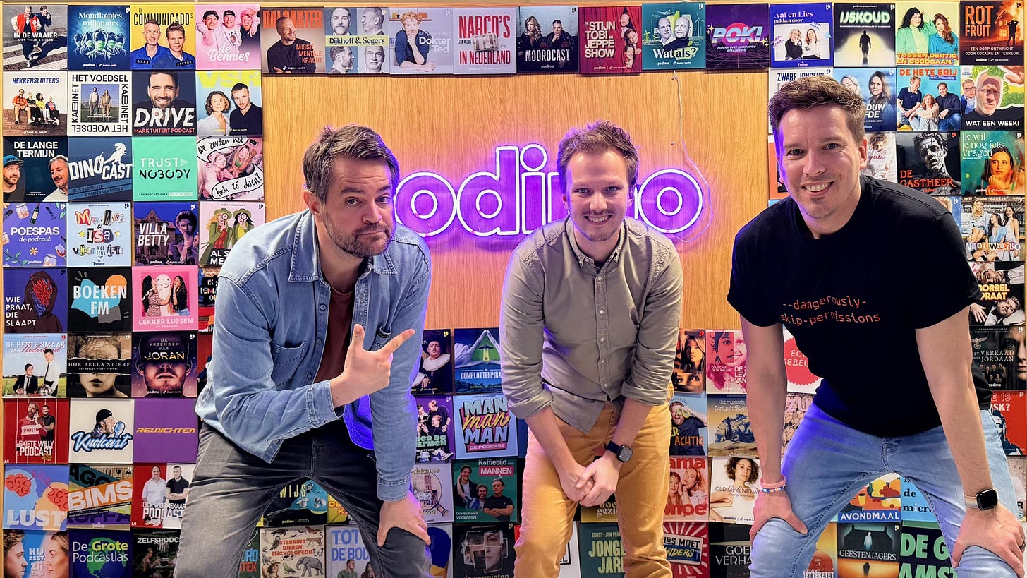 Promotiefoto voor de podcast Met Nerds om Tafel. Drie mannen poseren voor een wand bedekt met tientallen kleurrijke podcastcovers van het Nederlandse podcastnetwerk Podimo. Achter hen hangt een paars neonbord met het Podimo-logo. Van links naar rechts: Jurriaan Ubachs in een spijkerjasje, leunend naar voren met een uitgestoken vinger; Cas Burggraaf in een grijs overhemd, glimlachend met gevouwen handen; en Randal Peelen in een zwart t-shirt met de tekst "dangerously skip-permissions".