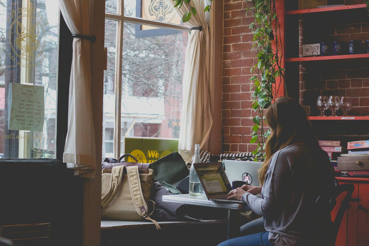 A woman sits at a small table in a cafe window, back to viewer, working on a laptop. A backpack and other personal items rest on a window seat opposite her.