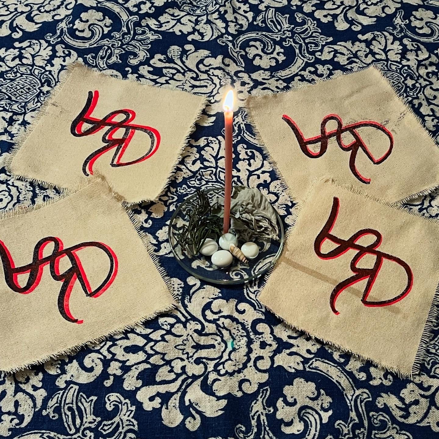 four fabric patches, printed with pink and purple sigils, surround a lit pink candle, herbs and precious stones