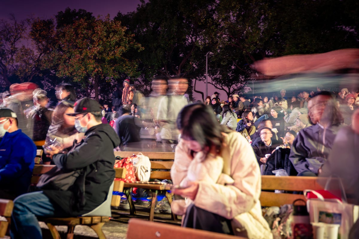 Crowds sit on benches and on the ground, and walk around in a park at night 