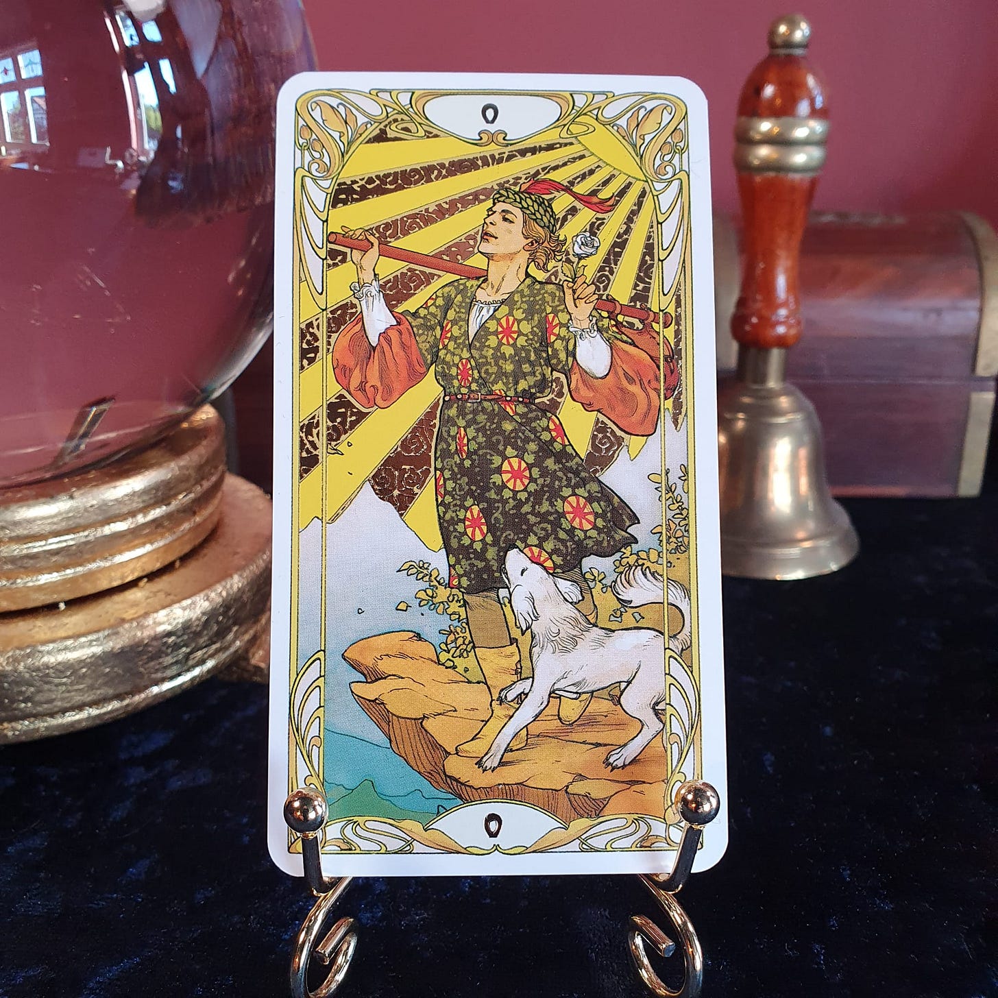 The Fool tarot card displayed on a gold stand against a red wall, used in the Tarot DMs interview with Elspeth Wilson, author of These Mortal Bodies.