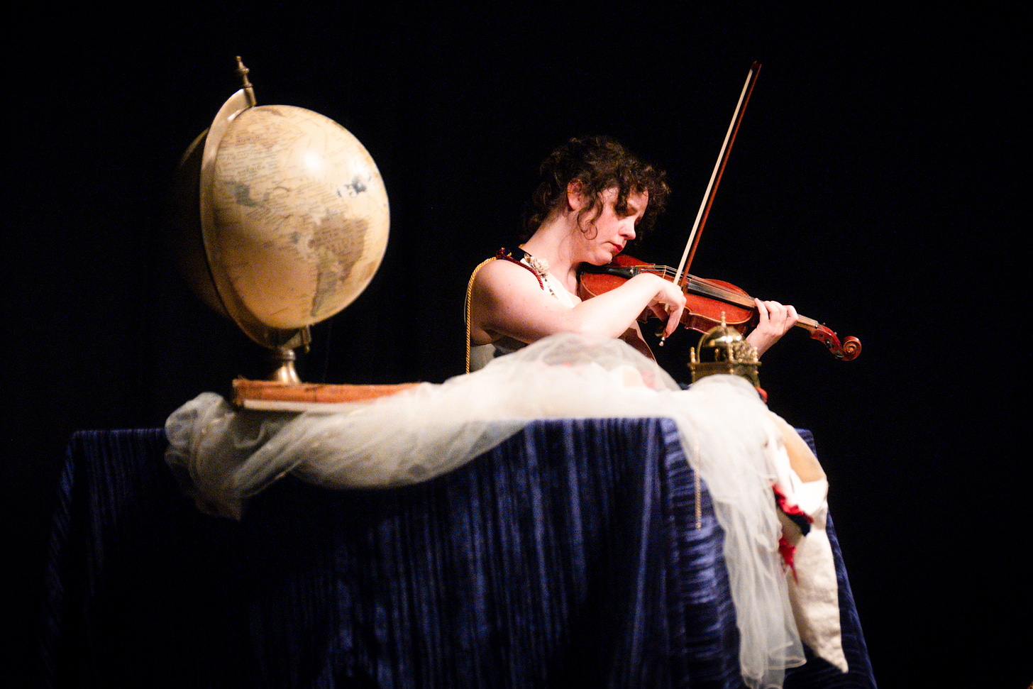 Tsarzi as Britannia plays violin pensively onstage - in the foreground, a table draped in blue velvet and set with a globe, a book of mythology, and a veil