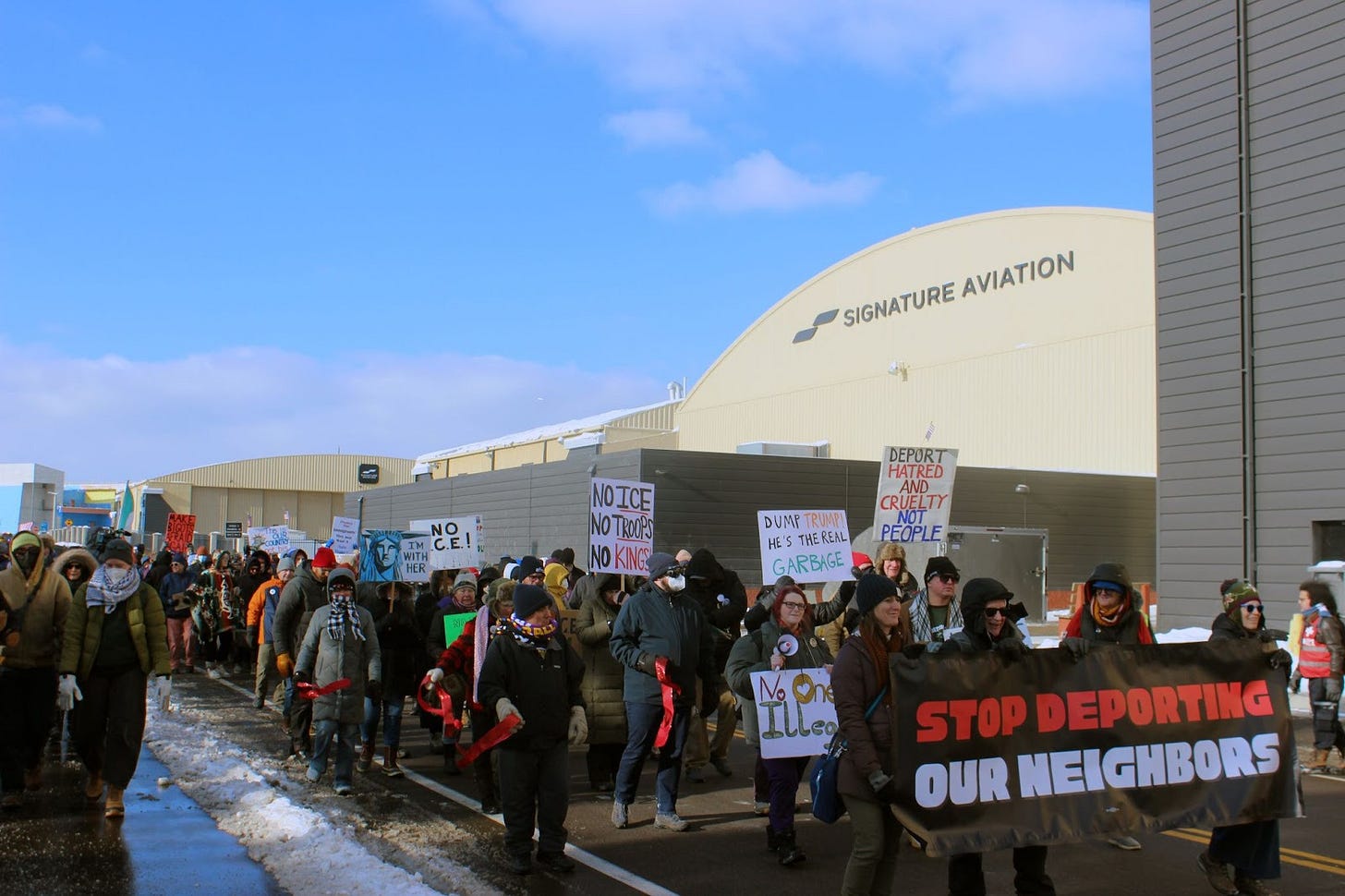 a crowd of people wearing winter gear march on the street holding signs and a banner that reads “stop deporting our neighbors” in front of a warehouse that reads “Signature Aviation"