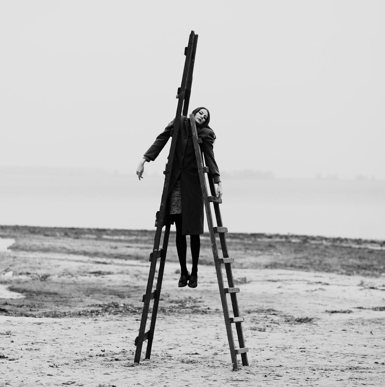 imagem é uma fotografia em preto e branco de uma mulher branca de cabelos escuros com roupas de frio – ela usa um casaco, uma saia, um cachecol longo, meia-calça e sapatos pretos, a paisagem atrás dela é de um campo arenoso. Na foto, a mulher tem cada um dos braços apoiados em longas escadas de madeiras que a suspendem do chão.