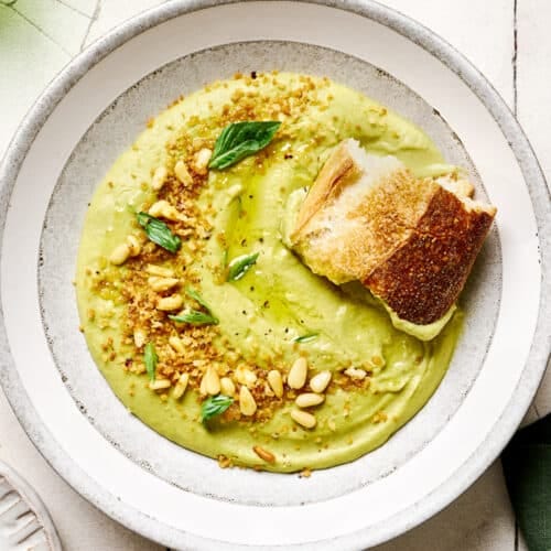 Hunk of sourdough in a bowl of creamy vegan broccoli soup in a white bowl on white tile table. Hunk of sourdough in a bowl of creamy vegan broccoli soup in a white bowl on white tile table.