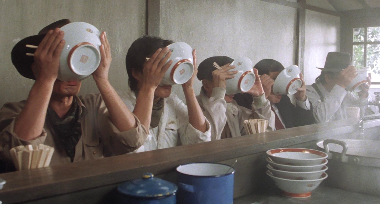 Five people sit at a counter, drinking ramen from bowls. Their hats and casual attire suggest a relaxed setting. The mood is lighthearted and communal.