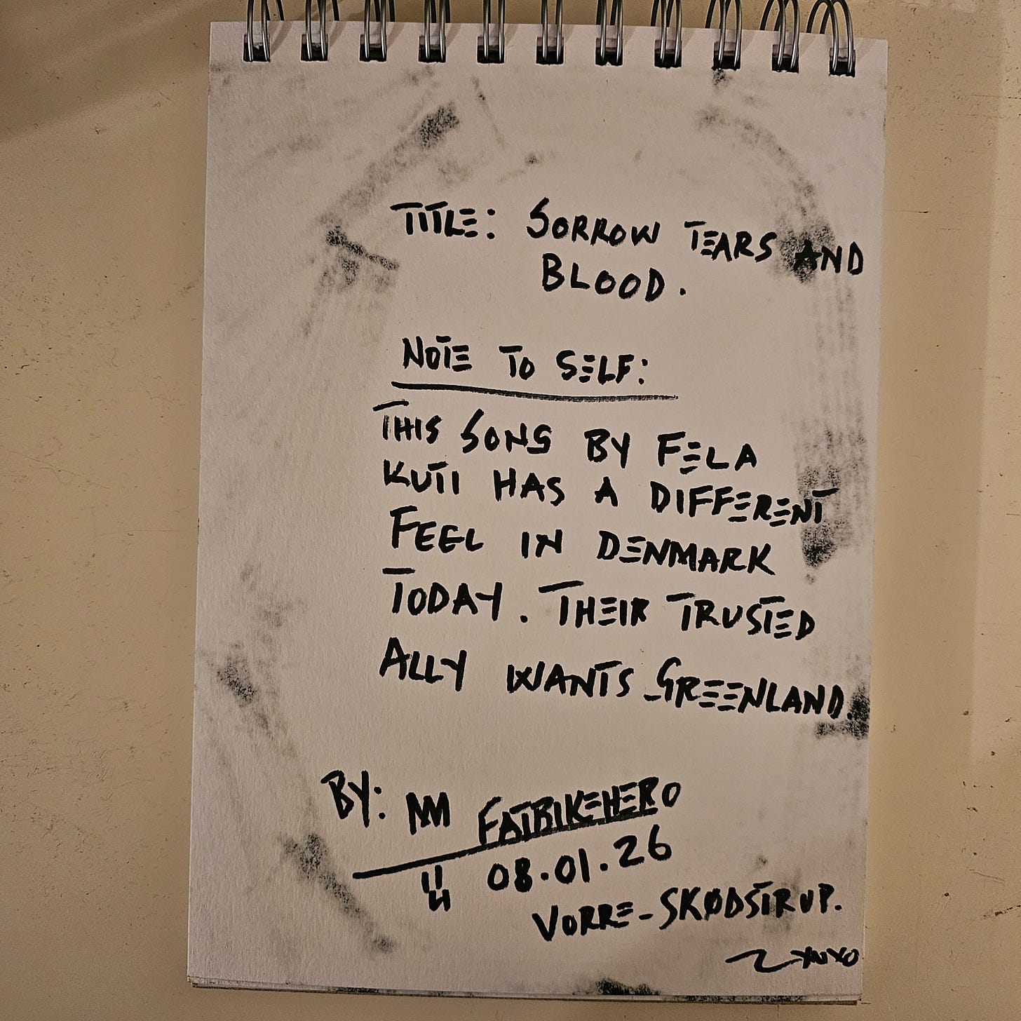 A photograph of a sketchbook page with handwritten text in black ink. The title reads "SORROW TEARS AND BLOOD." The note below states: "This song by Fela Kuti has a different feel in Denmark today. Their trusted ally wants Greenland." It is signed "By: M FatbikeHero," dated "08.01.26," and located in "Vorre - Skødstrup."