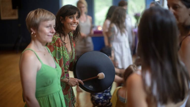 A smiling woman in a colorful dress plays a hand drum with a mallet during a group gathering, surrounded by people in conversation.