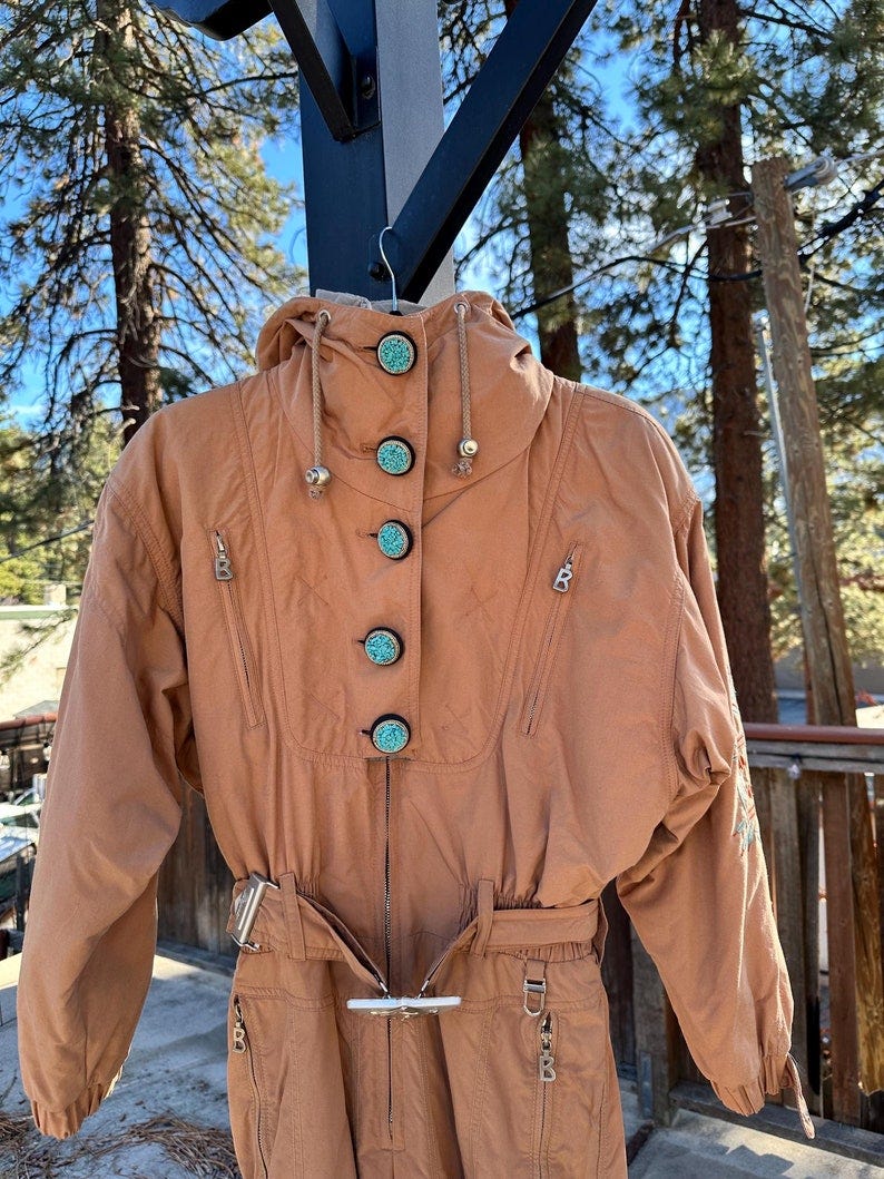 May include: Vintage women's ski suit in a light brown color. The suit features a high collar, a drawstring hood, and several pockets.  It has unique turquoise buttons and a belted waist.  The long sleeves and pants are designed for warmth and mobility on the slopes.  The suit shows some embroidered details on the sleeves.