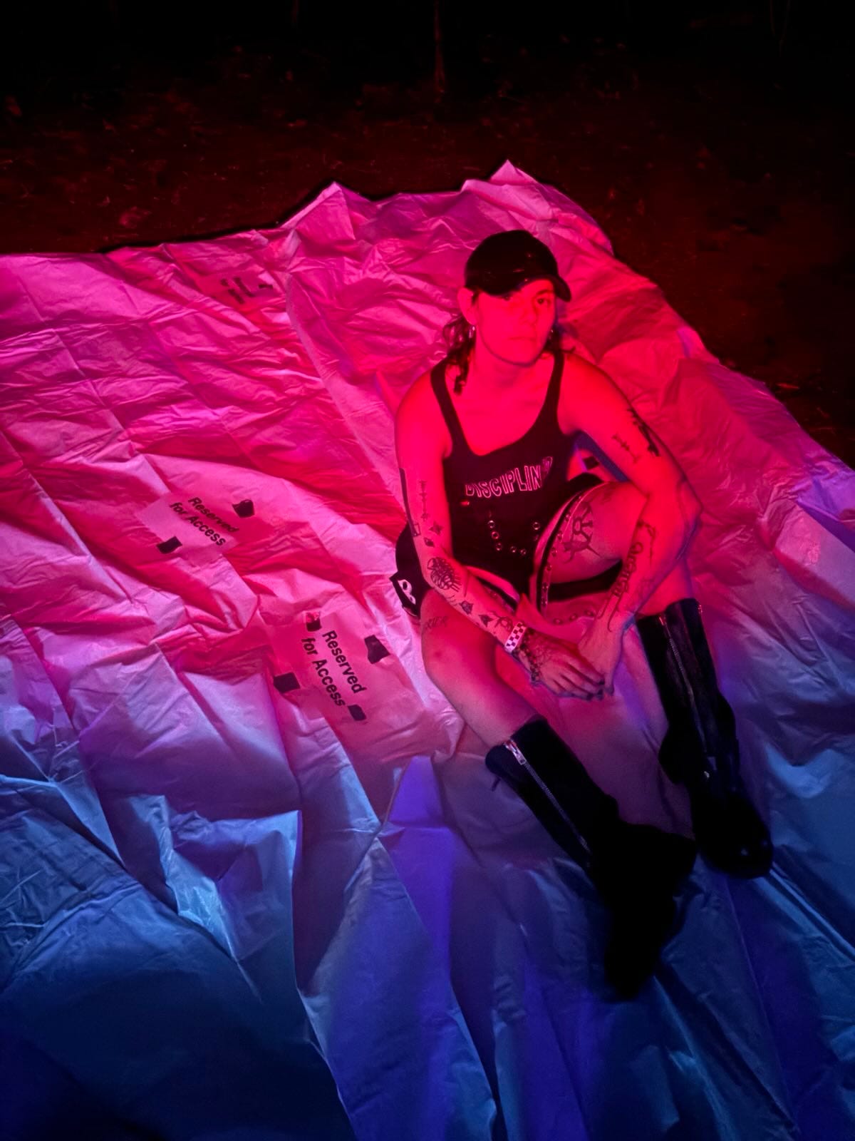 A nonbinary white person with tattoos in black clothing, including boots and a tank that says DISCIPLINE. They are bathed in a pink light sitting on a tarp with 2 pieces of paper that say RESERVED FOR ACCESS.