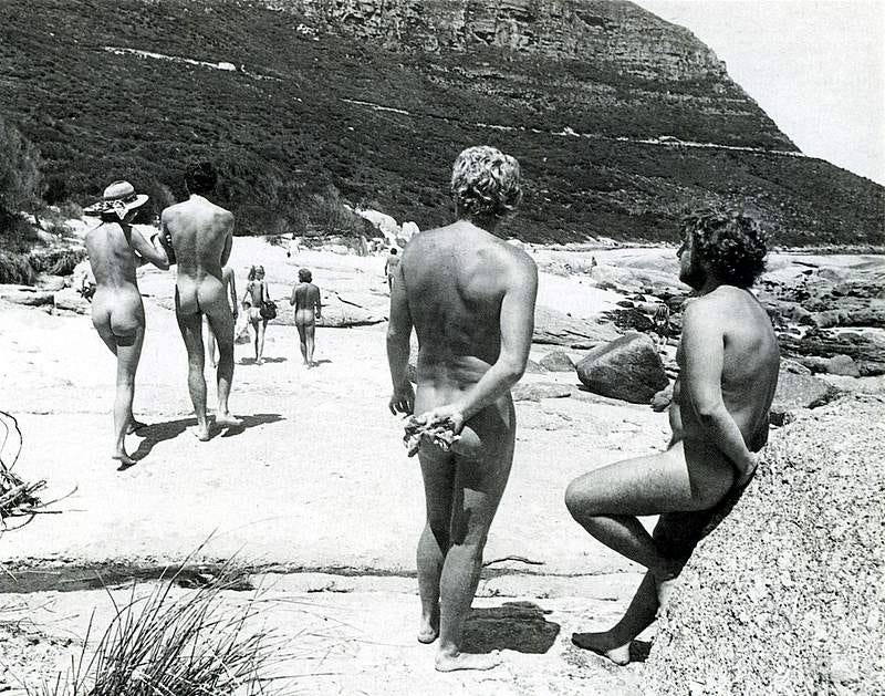 Black-and-white photo from the 1970s showing several nude adults walking and standing on Sandy Bay beach, with rocky shoreline and steep coastal cliffs in the background. Black-and-white photo from the 1970s showing several nude adults walking and standing on Sandy Bay beach, with rocky shoreline and steep coastal cliffs in the background.