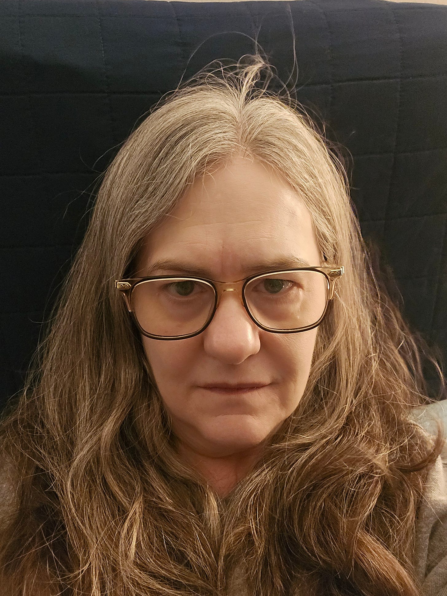 Close-up selfie of a woman with long hair and glasses, expression tired and serious. A visual representation of stress and overwhelm without digital enhancement.