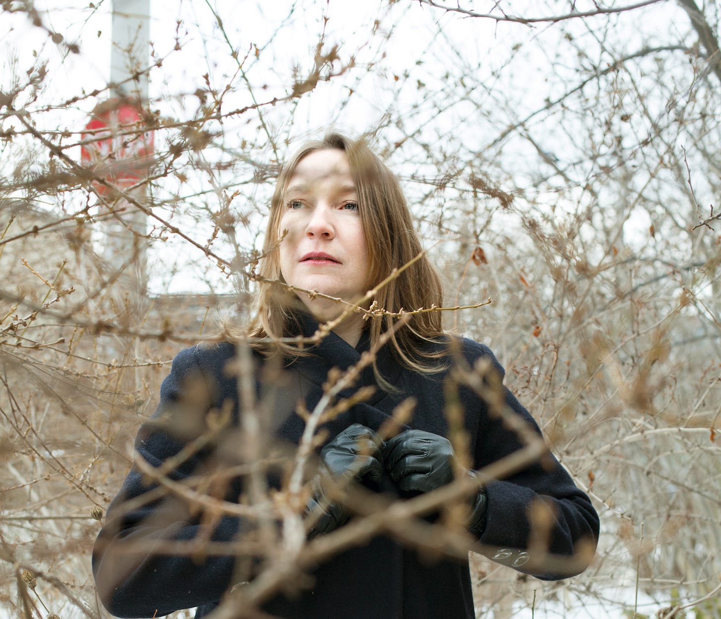 author sheila heti in a dark overcoat, standing behind branches