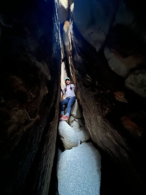 One row of three photos, left to right: Selfie photo of woman holding up her digital camera with sandy, gravelly driveway in the foreground and white one-story buildings in the background; Photo of woman standing in a darkened crevice, on boulders wedged between larger boulders and slabs of rock; Selfie photo of two women smiling atop a hill on a hike overlooking low lying hills that fall away to the Pacific Ocean.