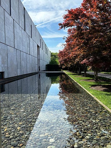 Modern building with stone facade next to a tree-lined reflecting pool. Clear sky above with scattered clouds, creating a serene atmosphere. Museums and wellbeing.