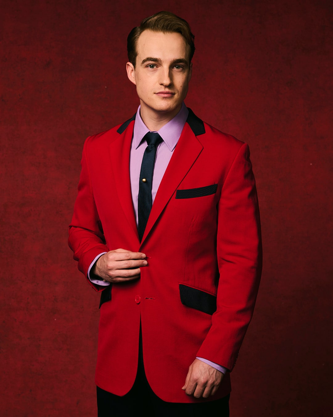 A man - Toby Miles - wearing a red blazer, purple shirt, and black tie against a red backdrop