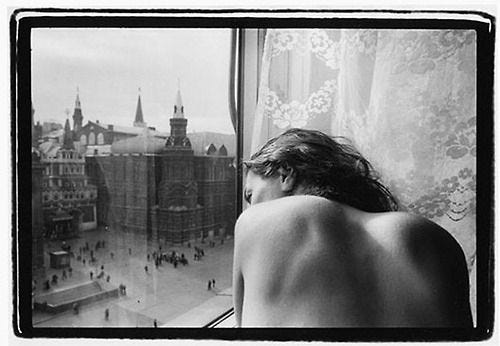 This may contain: black and white photograph of a woman looking out the window at an old cityscape