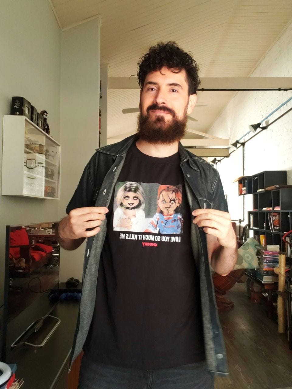 selfie colorida minha com uma camiseta estampada com Chucky e a noiva de chucky. A camisa é preta de estampa colorida, estou de calça jeans preta, barba grande escura e cabelos curtos cacheados, sorrindo. Por cima da camiseta há uma camisa jeans de manga curta aberta. O cenário é uma sala com televisão, luminárias e estante de livros.