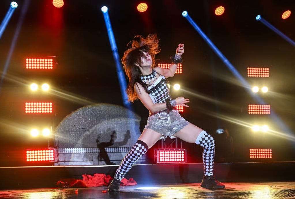 Performer dancing energetically on stage with bright lights and colorful beams. Wearing black and white checkered outfit, red cloth on floor.