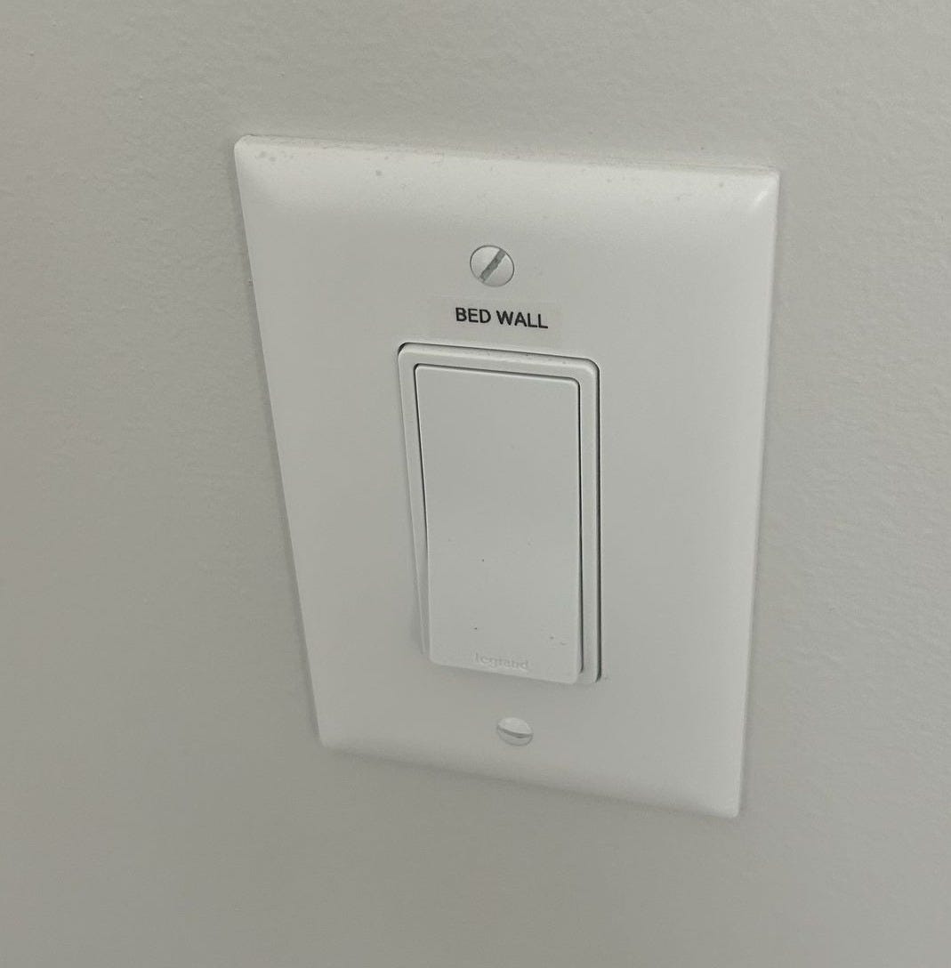 A white rocker light switch on a white wall, with the words BED WALL engraved vertically on the cover plate beside the switch. A white rocker light switch on a white wall, with the words BED WALL engraved vertically on the cover plate beside the switch.
