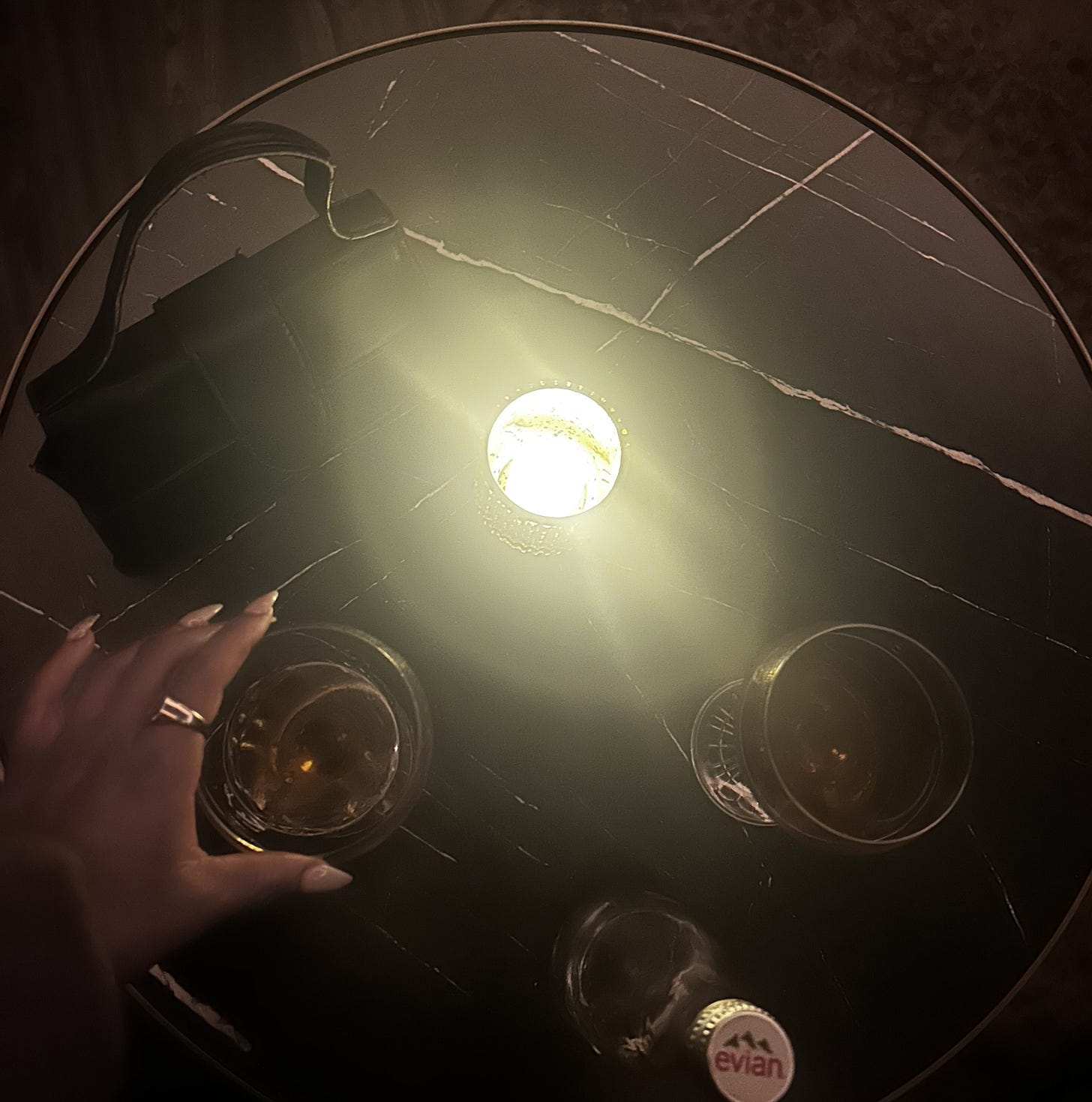 Top-down view of a small round marble table with a candle glowing at the center, two glasses of amber liquor, a hand reaching for one glass, and a small black purse resting beside the drinks. Top-down view of a small round marble table with a candle glowing at the center, two glasses of amber liquor, a hand reaching for one glass, and a small black purse resting beside the drinks.