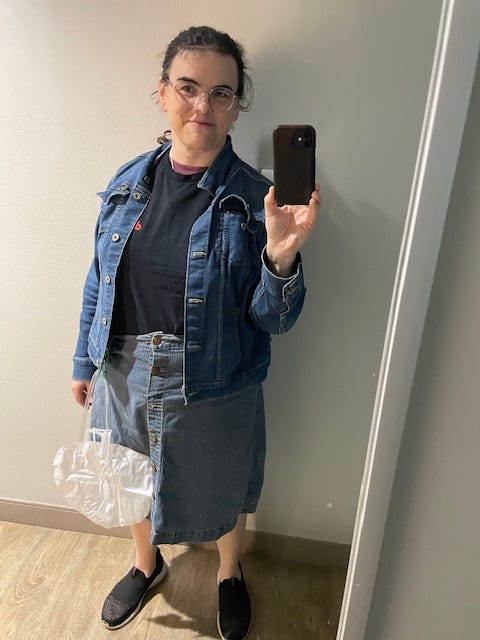 A woman taking a mirror selfie.  She's wearing a denim jacket, black t-shirt, and denim skirt.  A foley bag hangs from her skirt.