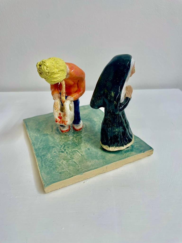A small handmade ceramic sculpture depicting two figures on a square green base. One figure, wearing an orange jumper and blue trousers, bends forward holding a pale object, while a second figure dressed in a long black religious habit stands beside them. A small handmade ceramic sculpture depicting two figures on a square green base. One figure, wearing an orange jumper and blue trousers, bends forward holding a pale object, while a second figure dressed in a long black religious habit stands beside them.